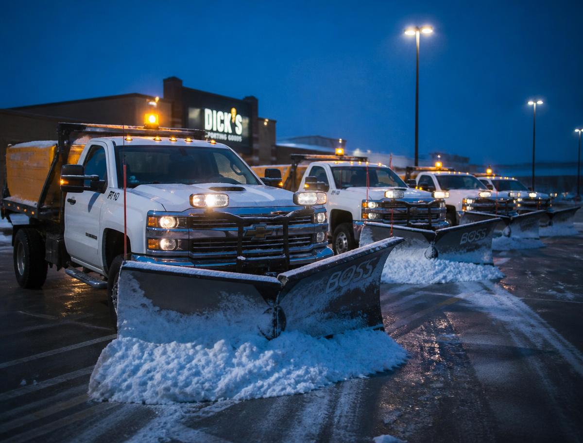 QSR-branded 3500-series commercial plow trucks with V-plows and tailgate salt spreaders staged in a lit commercial plaza lot